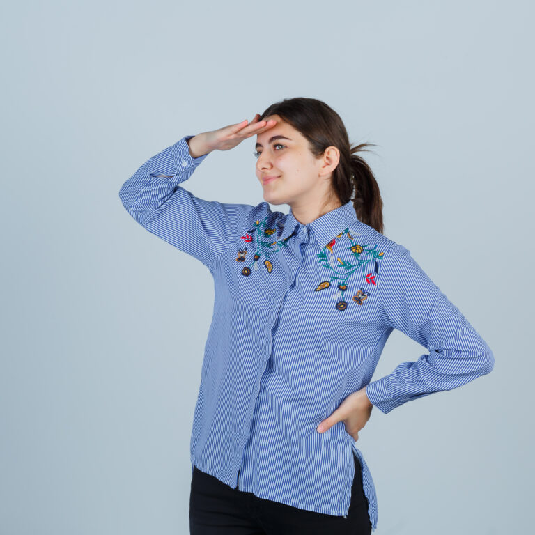 young lady holding hand on head to see clearly in shirt, pants and looking focused , front view.